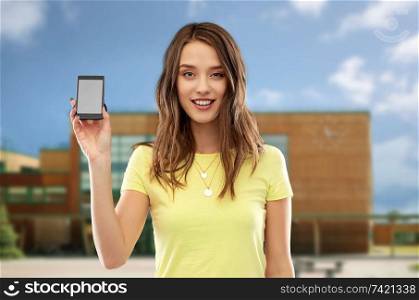 technology, education and people concept - smiling young woman or teenage girl in yellow t-shirt holding smartphone with blank screen over school background. teenage girl holding smartphone over school