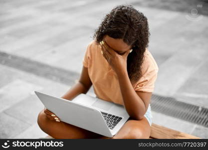technology, education and people concept - stressed african american student girl with laptop computer in city. stressed african student girl with laptop in city