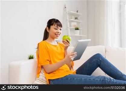 technology, education, diet and people concept - happy young asian woman sitting on sofa with tablet pc computer and green apple at home. happy asian woman with tablet pc and apple at home