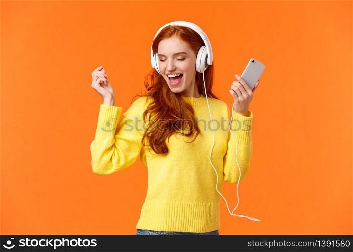 Technology, emotions and gadgets concept. Cheerful good-looking redhead woman dancing with hands up, closed eyes, smiling joyfully, enjoy awesome music sound in headphones, hold smartphone.. Technology, emotions and gadgets concept. Cheerful good-looking redhead woman dancing with hands up, closed eyes, smiling joyfully, enjoy awesome music sound in headphones, hold smartphone