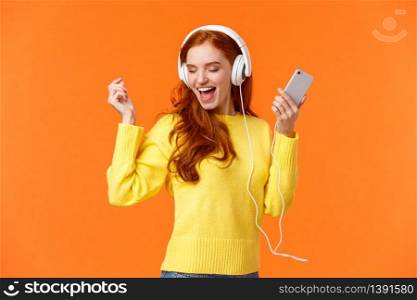 Technology, emotions and gadgets concept. Cheerful good-looking redhead woman dancing with hands up, closed eyes, smiling joyfully, enjoy awesome music sound in headphones, hold smartphone.. Technology, emotions and gadgets concept. Cheerful good-looking redhead woman dancing with hands up, closed eyes, smiling joyfully, enjoy awesome music sound in headphones, hold smartphone