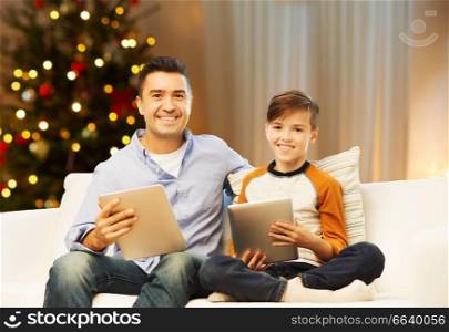 technology, family and holidays concept - happy father and son with tablet pc computer networking or playing at home over christmas tree lights background. father and son with tablet computers on christmas