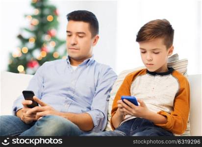 technology, family and people concept - father and son with smartphones texting message or playing game at home over christmas tree background. father and son with smartphones at christmas