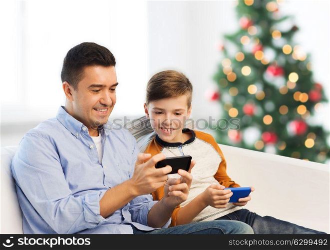 technology, family and people concept - happy father and son with smartphones texting message or playing game at home over christmas tree background. happy father and son with smartphones at christmas
