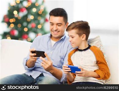 technology, family and people concept - happy father and son with smartphones texting message or playing game at home over christmas tree background. happy father and son with smartphones at christmas