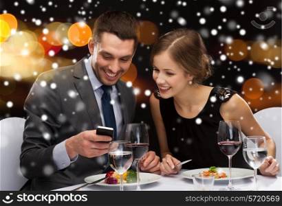 technology, food, christmas, holidays and people concept - smiling couple with smartphone eating at restaurant over night lights background