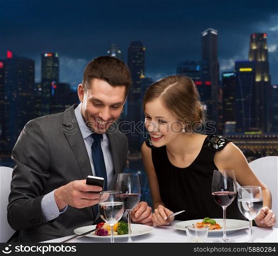 technology, food, holidays and people concept - smiling couple with smartphone eating at restaurant over night city background