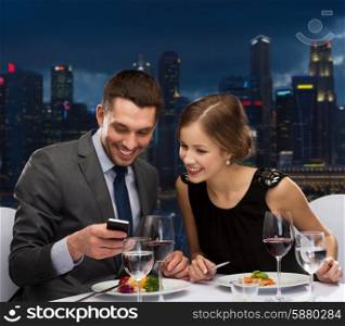 technology, food, holidays and people concept - smiling couple with smartphone eating at restaurant over night city background