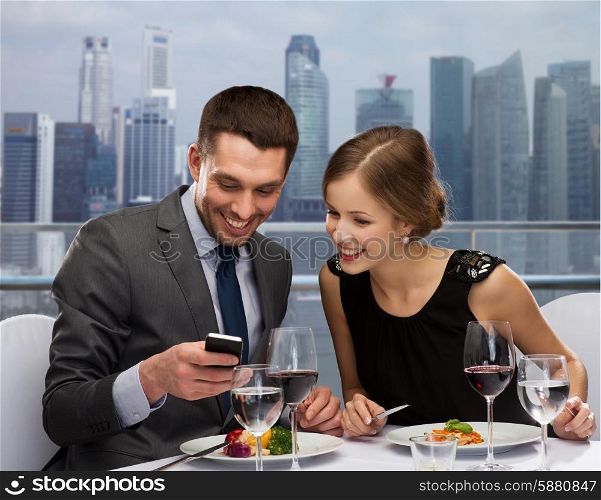 technology, food, holidays and people concept - smiling couple with smartphone eating at restaurant over city background
