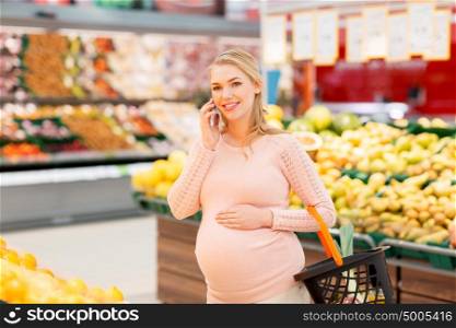 technology, food, pregnancy and people concept - happy pregnant woman with shopping basket calling on smartphone at grocery store or supermarket. pregnant woman with shopping basket and smartphone