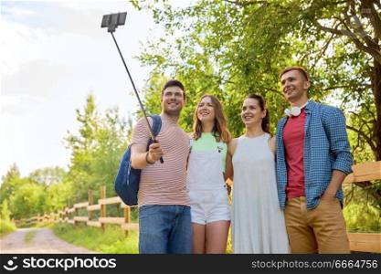 technology, friendship and hike concept - group of smiling friends with backpack taking picture by smartphone on selfie stick in summer. friends take picture by smartphone on selfie stick