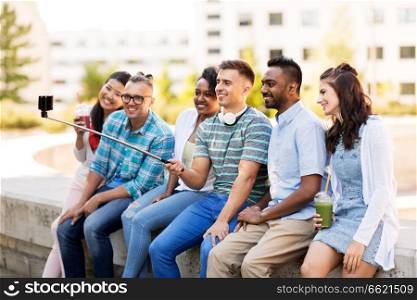 technology, friendship and international concept - group of students or friends with drinks taking picture by smartphone on selfie stick in city. friends taking picture by on selfie stick in city