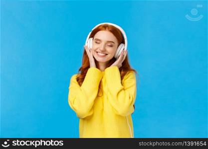 Technology, gadgets and lifestyle concept. Charming carefree redhead woman enjoy listening music in large white headphones, close eyes and smiling romantically, standing blue background.. Technology, gadgets and lifestyle concept. Charming carefree redhead woman enjoy listening music in large white headphones, close eyes and smiling romantically, standing blue background