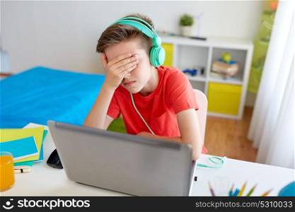 technology, gaming, cyberbullying and people concept - upset boy in headphones with laptop computer at home lost video game or suffer of bullying. boy in headphones playing video game on laptop