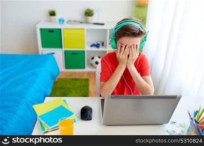 technology, gaming, cyberbullying and people concept - upset boy in headphones with laptop computer at home lost video game or suffer of bullying. boy in headphones playing video game on laptop