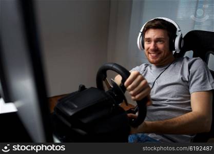 technology, gaming, entertainment and people concept - happy smiling young man in headphones with pc computer playing car racing video game at home and steering wheel
