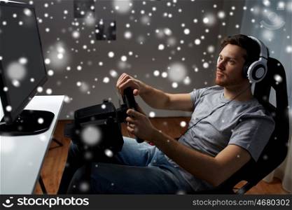 technology, gaming, entertainment and people concept - young man in headphones with pc computer playing car racing video game at home and steering wheel over snow