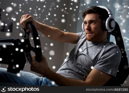technology, gaming, entertainment and people concept - young man in headphones with pc computer playing car racing video game at home and steering wheel over snow