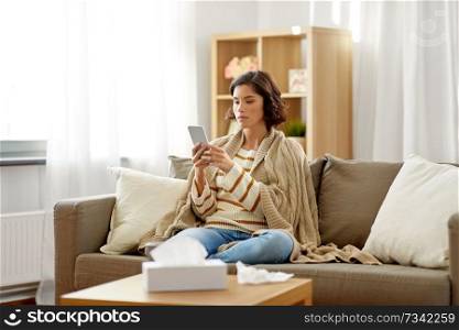technology, health and cold concept - sad sick woman in blanket using smartphone at home. sad sick woman in blanket using smartphone at home