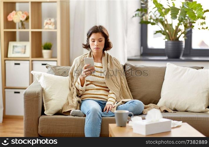 technology, health and cold concept - sad sick woman in blanket using smartphone at home. sad sick woman in blanket using smartphone at home