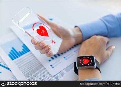 technology, health care and people concept - close up of woman hand holding transparent smartphone and smartwatch with heart icon at office