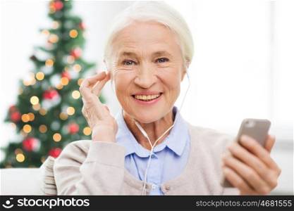 technology, holidays age and people concept - happy senior woman with smartphone and earphones listening to music at home over christmas tree background
