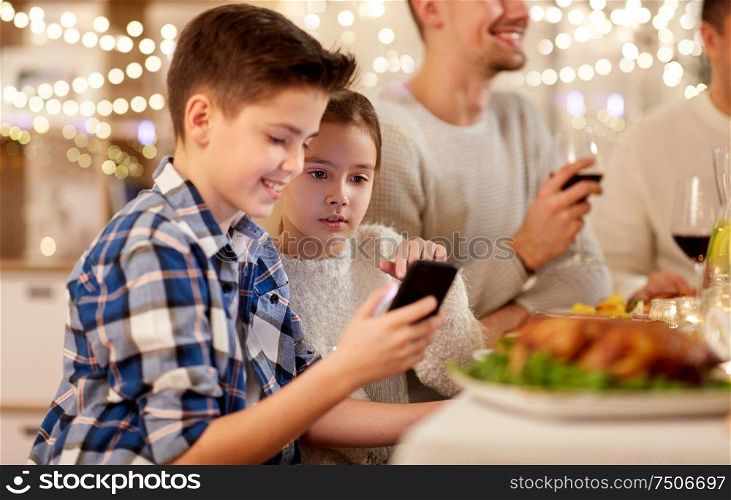 technology, holidays and people concept - happy boy with sister using smartphone having family dinner party at home. boy with sister using smartphone at family dinner