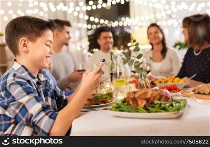 technology, holidays and people concept - happy boy with smartphone having family dinner party at home. boy with smartphone at family dinner party