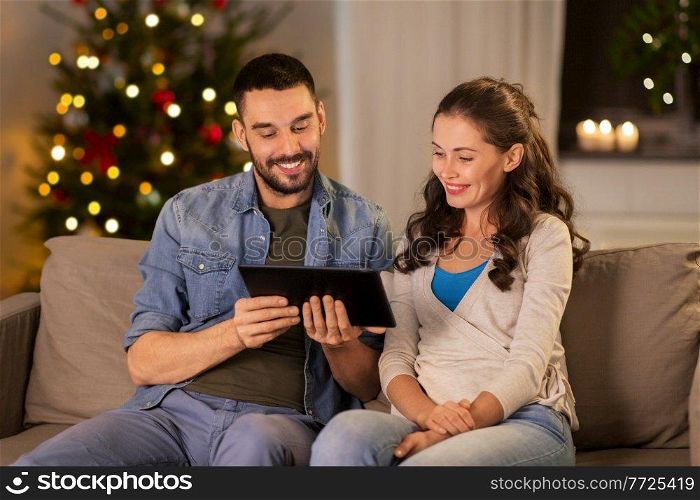 technology, holidays and people concept - happy couple using tablet computer at home in evening over christmas tree lights on background. happy couple using tablet pc at home on christmas