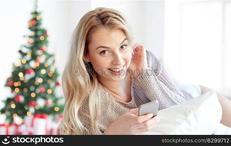 technology, holidays and people concept - happy young woman lying in bed with smartphone at home bedroom over christmas tree background. happy woman with smartphone in bed at christmas
