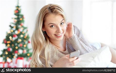 technology, holidays and people concept - happy young woman lying in bed with smartphone at home bedroom over christmas tree background. happy woman with smartphone in bed at christmas