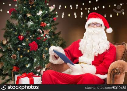 technology, holidays and people concept - man in costume of santa claus with tablet pc computer, gift and christmas tree sitting in armchair over garland lights background. santa claus with tablet pc and christmas tree