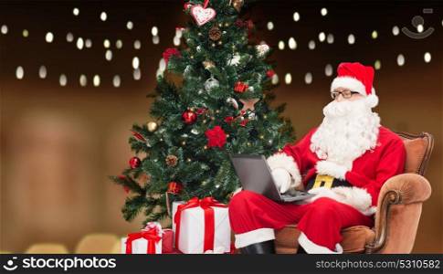 technology, holidays and people concept - man in costume of santa claus with laptop computer, gifts and christmas tree sitting in armchair over garland lights background. man in costume of santa claus with laptop