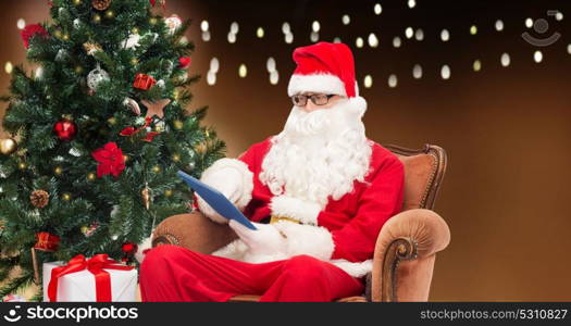 technology, holidays and people concept - man in costume of santa claus with tablet pc computer, gift and christmas tree sitting in armchair over garland lights background. santa claus with tablet pc and christmas tree