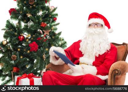 technology, holidays and people concept - man in costume of santa claus with tablet pc computer, gifts and christmas tree sitting in armchair