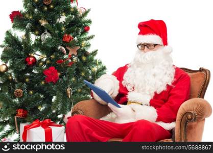 technology, holidays and people concept - man in costume of santa claus with tablet pc computer, gifts and christmas tree sitting in armchair