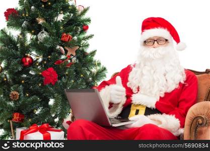technology, holidays and people concept - man in costume of santa claus with laptop computer, gifts and christmas tree sitting in armchair