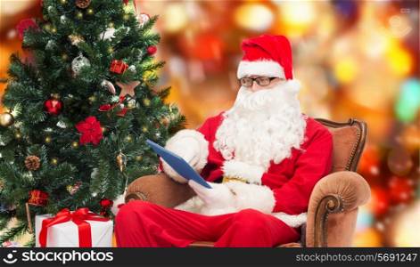 technology, holidays and people concept - man in costume of santa claus with tablet pc computer, gifts and christmas tree sitting in armchair over red lights background