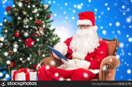 technology, holidays and people concept - man in costume of santa claus with tablet pc computer, gifts and christmas tree sitting in armchair over blue snowy background