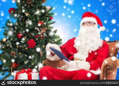 technology, holidays and people concept - man in costume of santa claus with tablet pc computer, gifts and christmas tree sitting in armchair over blue snowy background