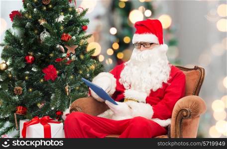 technology, holidays and people concept - man in costume of santa claus with tablet pc computer, gifts and christmas tree sitting in armchair over lights background