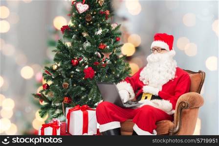 technology, holidays and people concept - man in costume of santa claus with laptop computer, gifts and christmas tree sitting in armchair over lights background