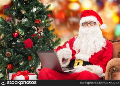 technology, holidays and people concept - man in costume of santa claus with laptop computer, gifts and christmas tree sitting in armchair over red lights background