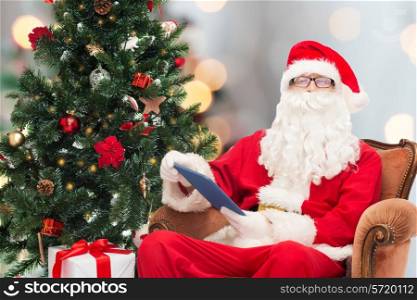 technology, holidays and people concept - man in costume of santa claus with tablet pc computer, gifts and christmas tree sitting in armchair over lights background
