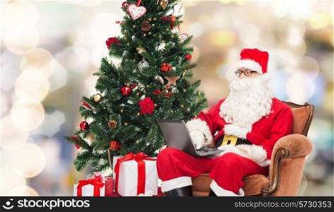 technology, holidays and people concept - man in costume of santa claus with laptop computer, gifts and christmas tree sitting in armchair over lights background