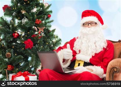 technology, holidays and people concept - man in costume of santa claus with laptop computer, gifts and christmas tree sitting in armchair over blue lights background