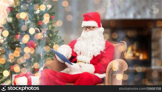 technology, holidays and people concept - man in costume of santa claus with tablet pc computer, gifts and christmas tree sitting in armchair over home fireplace and lights background
