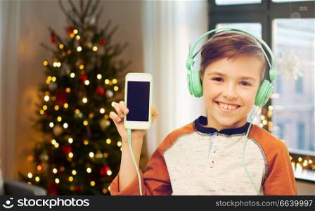 technology, holidays and people concept - smiling boy with smartphone and headphones listening to music at home over christmas tree lights on background. boy with smartphone and headphones on christmas
