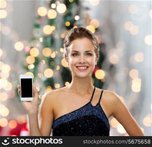technology, holidays and people concept - smiling woman in evening dress showing smartphone blank screen over christmas tree and gifts background
