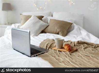 technology, holidays, christmas and winter concept - cozy bedroom with laptop computer, coffee cup and croissant on bed at cozy home